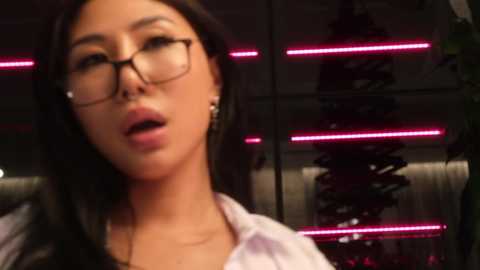 Media: Video of an Asian woman with long black hair, glasses, and light skin, wearing a white blouse, with a dimly lit background featuring red neon lights and metallic structures.