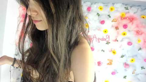 Media: Video of a young woman with long, wavy brown hair, wearing a black top, standing in front of a floral wall backdrop.