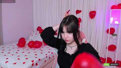 Media: A young woman with pale skin, dark hair, and a black dress stands in a bedroom with red balloons and a bed covered in hearts, appearing upset.