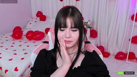 Media: A video of a young woman with long black hair, wearing a black top, seated on a pink gaming chair in a room decorated with red balloons and heart-shaped decorations.