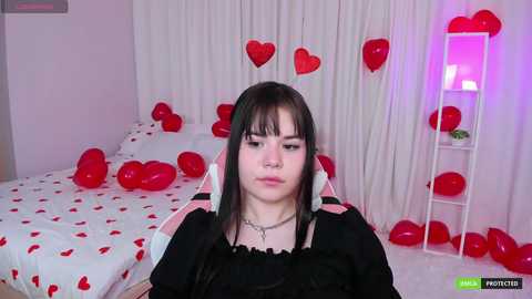 Media: A video of a young woman with long, straight black hair, wearing a black dress, seated in a room adorned with red heart decorations and balloons.