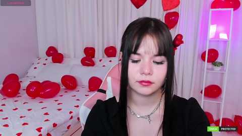 Media: Video of a young Asian woman with straight black hair, wearing red lipstick, black jacket, and silver necklace, surrounded by red balloons and rose petals in a bedroom.