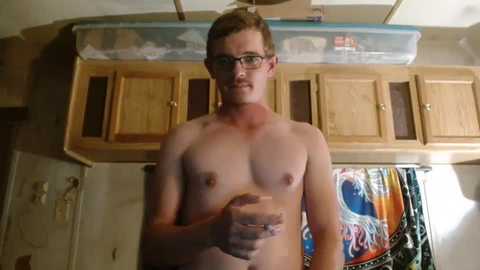 Media: Video of a shirtless, light-skinned man with short brown hair, glasses, and a slight beard, standing indoors near a wooden cabinet and a surfboard poster.