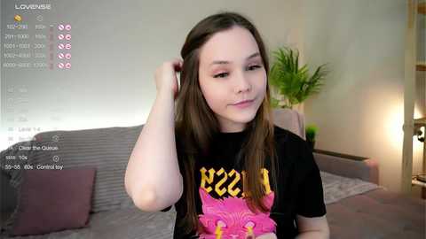 Media: Video of a young woman with fair skin and long brown hair, wearing a black t-shirt with a pink design. She is seated in a modern living room with a grey couch, green plants, and a lamp.