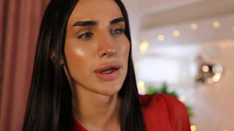 Video of a woman with long, straight black hair, fair skin, and full lips, wearing a sheer red top, speaking with a soft expression. Background features blurred, warm-toned lights and a hint of greenery.