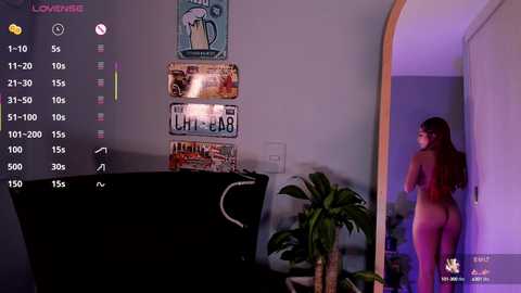 Media: Video of a nude woman with long brown hair, viewed from behind, standing in a dimly lit room with posters and a potted plant.