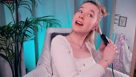 Media: Video of a young blonde woman with fair skin, wearing a white cardigan, sitting on a chair, using a hairdryer. Background includes potted plants, a towel hanging, and a framed picture.