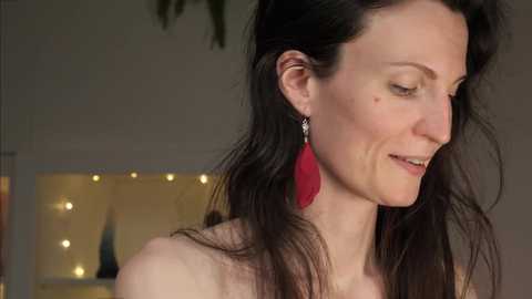 Media: A video of a smiling, light-skinned woman with long, dark hair, wearing red, dangling earrings, standing indoors with a blurred background featuring a wall-mounted, illuminated abstract art piece.