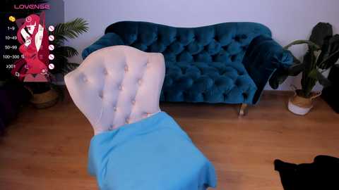 Media: A video shows a teal velvet chaise lounge with a white cushion, draped in a blue blanket, in a modern room with wooden floors, potted plants, and a pink cartoon overlay.