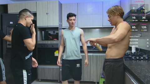 Video of three shirtless men in a modern kitchen. One man, wearing shorts, holds a phone, while another, wearing a tank top, stands in the center. A third man, shirtless, gestures with a knife on a counter.