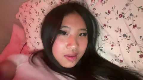 Media: Video of an East Asian woman with long black hair, light skin, and a septum piercing, lying on a floral-patterned pillow. She has a soft, dreamy expression.