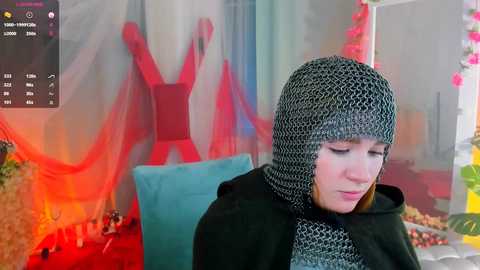 Media: Video of a fair-skinned woman in a medieval-style chainmail hood, standing against a backdrop of a red-clad mannequin, with a digital interface on the left showing streaming controls.