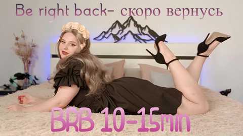 Media: Video of a curvy, fair-skinned woman with long, wavy brown hair, wearing a black dress and black high heels, lying on a beige bed with a mountain art backdrop. Text overlays read, \"Be right back - 10 minutes\" and \"BBB-10-15min.\