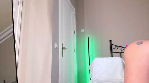 Media: Video of a beige-walled bedroom with a white door, a green-tinted LED strip, and a black wrought-iron bed with white bedding.
