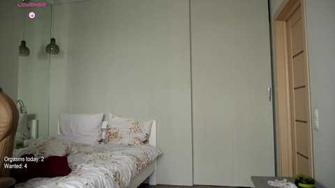 Media: Video of a minimalist, light-colored bedroom with a double bed, floral bedding, white walls, and a closed wardrobe.