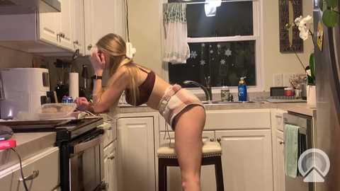 Media: Video of a blonde woman in lingerie, bent over a kitchen counter, with white cabinets and a window in the background.