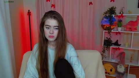 Media: Video of a young woman with long brown hair, wearing a light blue sweater, sitting on a beige couch. Background includes a red-lit wall, a white shelf with toys and a colorful fish tank, and pink curtains.