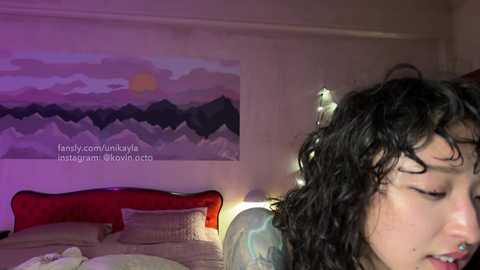 Media: A video of a young woman with curly dark hair and a nose ring, lying on a bed with a red headboard, in a dimly lit room with a mountain sunset painting on the wall.