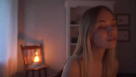 Media: Video of a blonde woman with straight hair, fair skin, and a slim physique, wearing a sleeveless top, smiling softly. The background features a dimly lit room with a wooden chair, a lit candle, and a bookshelf with framed pictures.