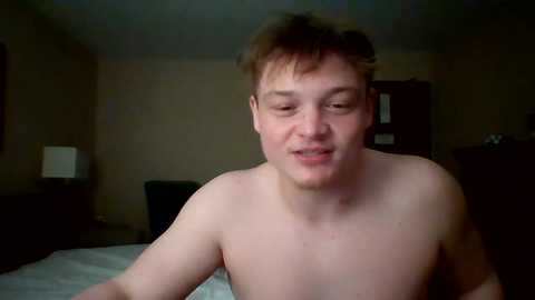 Media: Video of a young, pale-skinned, short-haired man with a slight smile, shirtless, in a dimly lit bedroom with beige walls and a dark wooden dresser.