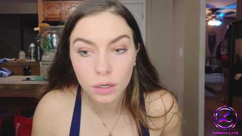 Media: Video of a young woman with fair skin, long brown hair, wearing a blue apron, in a kitchen with wooden cabinets, counter clutter, and ceiling fan.