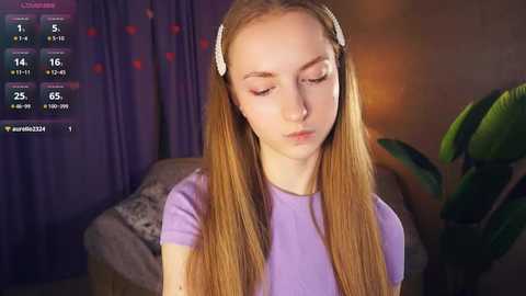 Media: Video of a young Caucasian girl with long blonde hair, wearing a lavender top, sitting indoors, with a virtual weather app displaying rain and a temperature of 25\u00b0C.