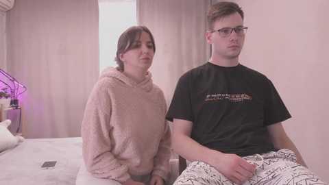 Media: A video of two people in a bedroom: a woman with light skin, dark hair, wearing a light pink fuzzy hoodie, and a man with glasses, light skin, short brown hair, wearing a black t-shirt and patterned pajama pants.