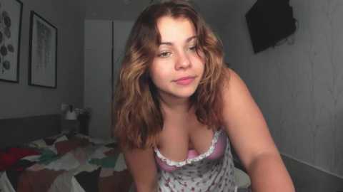 Media: Video of a young Caucasian woman with wavy brown hair, wearing a low-cut floral camisole, leaning forward, in a dimly lit bedroom with a bed, framed art, and a mounted TV on the wall.