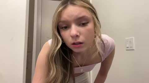 Media: Video of a young Caucasian woman with long, blonde hair, wearing a light pink crop top, leaning forward, looking down, in a minimalistic room with white walls and a light switch.