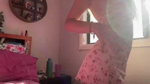 Media: Video of a woman in a floral dress adjusting her bra, standing near a window in a cozy, pink-themed bedroom with a wooden bed, a round mirror, and decorative items on a shelf.