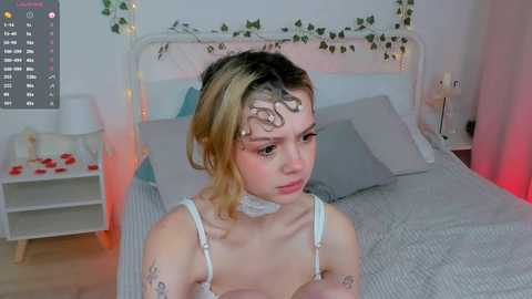 Media: Video of a young woman with blonde hair, wearing a white choker and white lingerie, sitting on a bed with grey bedding in a dimly lit, cozy room.