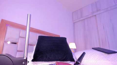 Media: Video of a minimalist bedroom with a black throw pillow on a white bed, wooden headboard, beige curtains, and a black keyboard on the right side.
