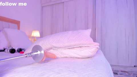 Media: Video of a bed with white sheets, a white duvet, and a man's erect penis protruding from beneath the duvet. The room has white walls, a wooden headboard, and a bedside lamp.