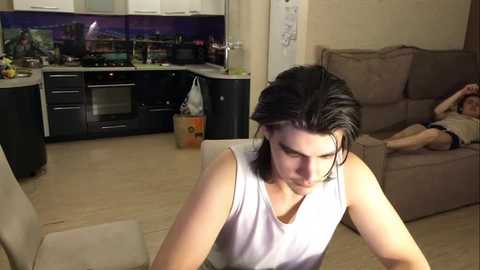 Media: Video of a young man with wet hair, wearing a white tank top, sitting on a beige chair in a cozy living room with a beige couch, kitchen in the background.
