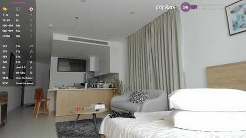 Media: Video of a modern, minimalist bedroom with a beige sofa, white bedding, light wooden headboard, and beige curtains. A small kitchenette is visible in the background.