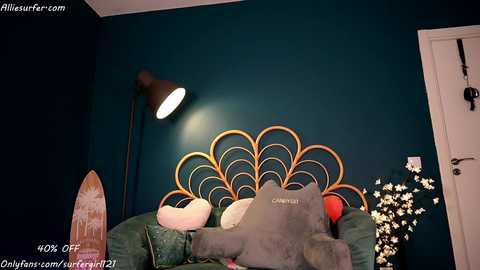 Media: Video of a cozy bedroom with teal walls, a wooden headboard with a peacock design, plush toys, a tall lamp, and a surfboard.