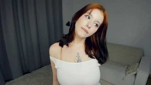 Media: Video of a fair-skinned woman with shoulder-length, dark brown hair, wearing a white off-shoulder top, sitting on a beige couch. She has a tattoo on her chest and is looking upward thoughtfully.