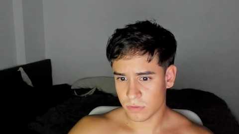 Media: Video of a shirtless young man with short, dark hair, fair skin, and a serious expression. He sits on a bed with black sheets and pillows in a dimly lit room with plain white walls.