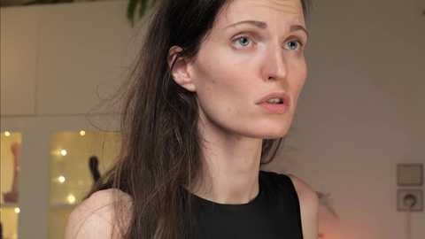 Media: Video of a fair-skinned woman with blue eyes and long dark hair, wearing a black sleeveless top, indoors with blurred background lights.