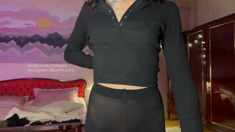 Media: Video of a person in a black sheer top and matching sheer skirt, standing in a bedroom with a red tufted headboard, a mountain mural on the wall, and a mirrored closet.