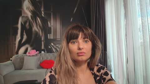 Media: Video of a young woman with straight, shoulder-length brown hair, wearing a leopard print top, sitting indoors. Background includes a grey sofa, black and white art print, and sheer white curtains.