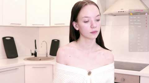 Media: Video of a young Caucasian woman with fair skin, long black hair, wearing a white off-shoulder sweater, standing in a modern kitchen with white cabinets, a stainless steel sink, and black speakers.