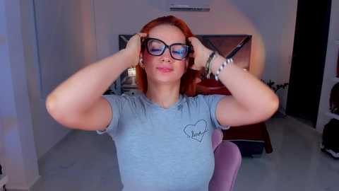 Media: Video of a light-skinned woman with red hair, wearing black-rimmed glasses, blue t-shirt with heart design, and white bracelet, adjusting her hair in a modern, well-lit room with dark furniture.