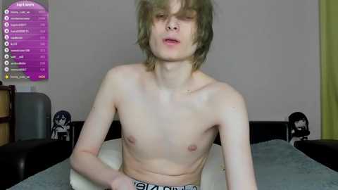 Video of a young, slender, shirtless, pale-skinned boy with messy hair, sitting at a table. Background features a purple menu screen, a white wall with cartoon figures, and a green curtain.