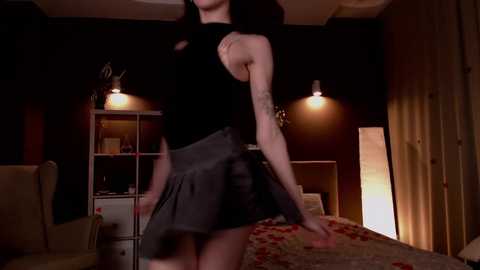 Media: A video of a slender, light-skinned woman in a black sleeveless top and gray pleated skirt dancing in a dimly lit bedroom with dark walls, a white shelf, and a floral-patterned bedspread.