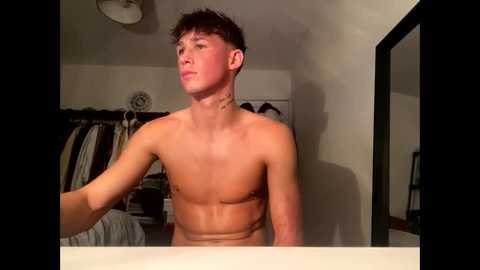Media: Video of a shirtless, lean young man with short brown hair, standing in a dimly lit bedroom with a mirror and open closet filled with clothes in the background.