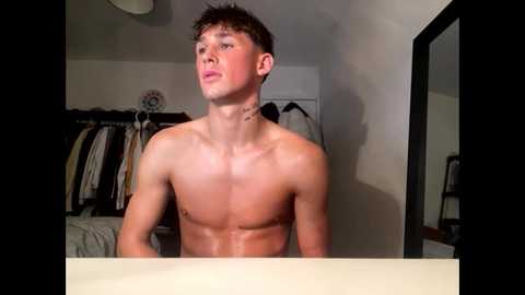 Media: Video of a shirtless, lean, young man with short brown hair, standing in a dimly lit bedroom with a cluttered closet in the background.