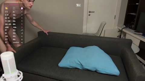 Media: A video of a naked woman with a slim physique and small breasts, adjusting a blue pillow on a gray couch in a modern, minimalist living room.