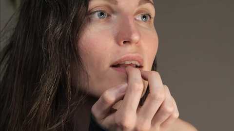 Media: A close-up video of a fair-skinned woman with long, dark brown hair and blue eyes, wearing a black top. She is biting her lower lip thoughtfully, with her hands near her mouth. The background is a neutral, blurred beige.