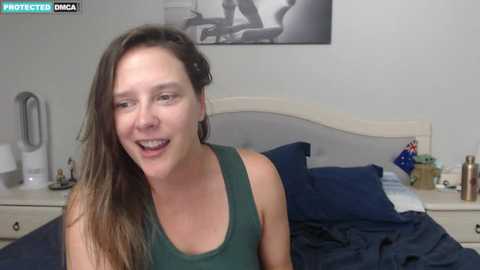 Media: Video of a smiling, light-skinned woman with long, wavy brown hair, wearing a green tank top, sitting on a bed with navy sheets and pillows.